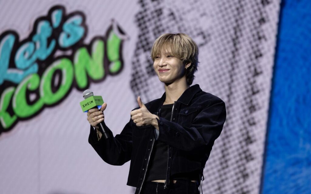 Taemin onstage holds a microphone and gives a thumbs-up.