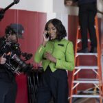 A woman in a green jacket and black pants standing near a cameraman with a cell phone to her ear.