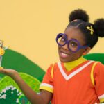 A girl in a purple glasses and an orange top holds her hand out with a yellow robot.