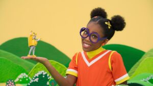 A girl in a purple glasses and an orange top holds her hand out with a yellow robot.