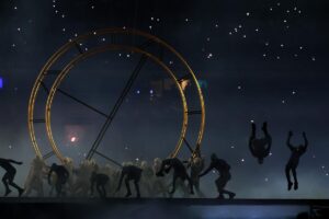 People in silhouette moving in front of a giant, gold-colored ring.