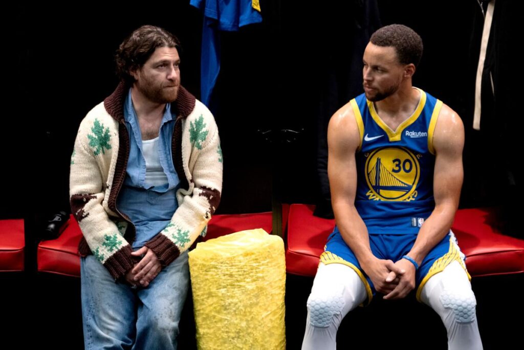 A man in a white and brown sweater sits next to Steph Curry in a blue and yellow basketball uniform.