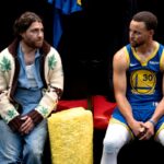 A man in a white and brown sweater sits next to Steph Curry in a blue and yellow basketball uniform.