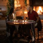 A woman and man sitting a table with a fire burning in a fireplace behind them.