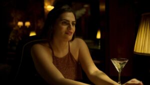 A woman in a spaghetti-strap dress sits at a restaurant table with a drink in front of her.