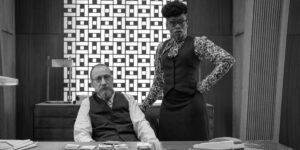 A black and white image of a man seated at an office desk with a woman standing next to him.