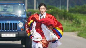 A woman wearing a colorful and not contemproary red, yellow, and blue dress runs on a highway with a jeep behind her in Mr. Plankton