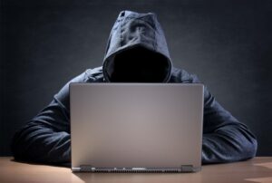 A hacker wearing a hoodie sits in front of an open laptop.