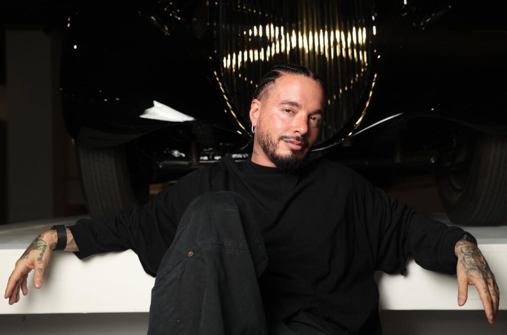 J Balvin in a black outfit poses in front of a car grill