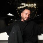 J Balvin in a black outfit poses in front of a car grill