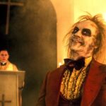 Beetlejuice in a red tux holding out his hand with a preist behind a podium in the background in Beetlejuice Beetlejuice