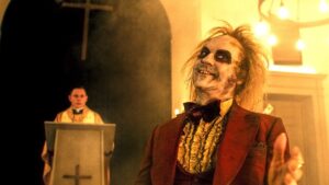 Beetlejuice in a red tux holding out his hand with a preist behind a podium in the background in Beetlejuice Beetlejuice