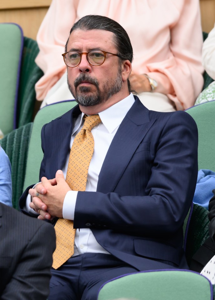 Dave Grohl wearing a navy suit with a white shirt and yellow tie for the annual tennis championship