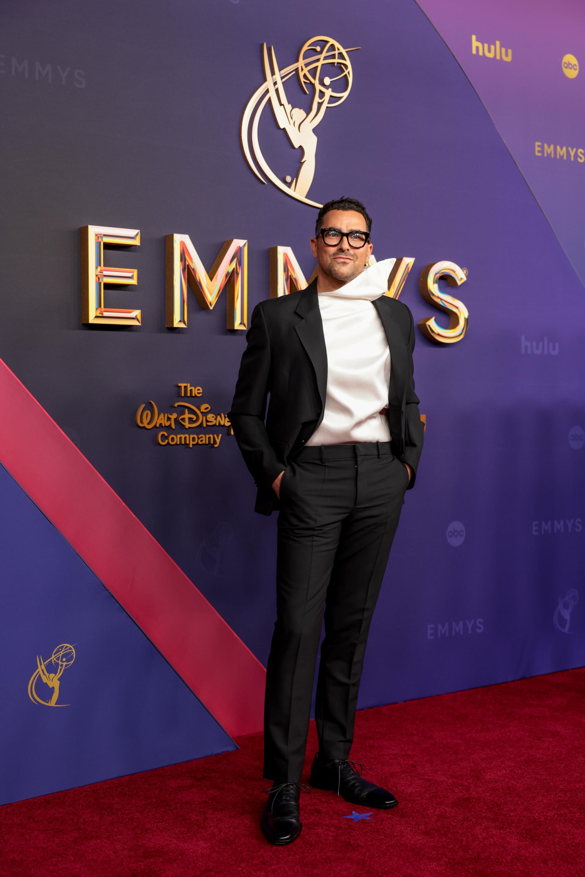 Dan Levy wears a white shirt with an exaggerated collar and a black suit. 