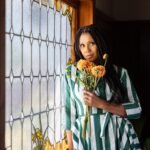 A woman holding flowers leans against the frame of a stained-glass window.