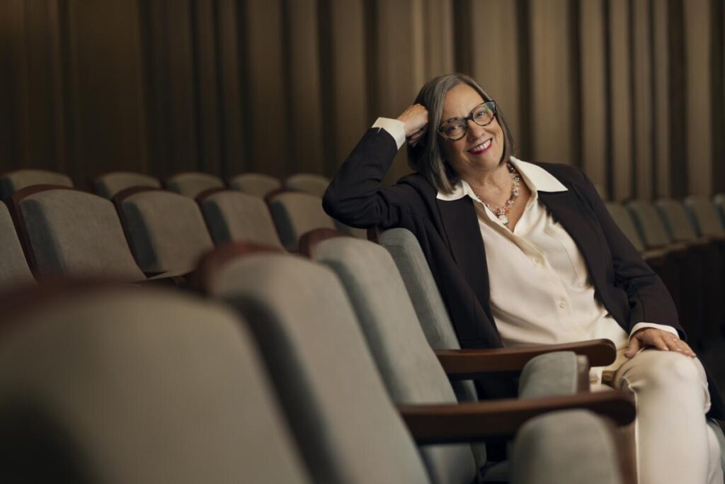 A smiling woman in glasses relaxes on a muted blue-green seat  inside an auditorium.