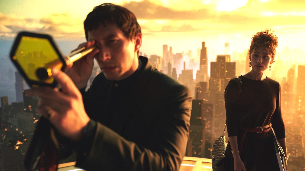 Adam Driver looks through a scope into a colored glass while Nathalie Emmanuel stands behind him high above the city in Megalopolis