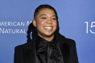 Punkie Johnson with braids wearing a black suit jacket and a black silk blouse. She poses in front of a blue backdrop