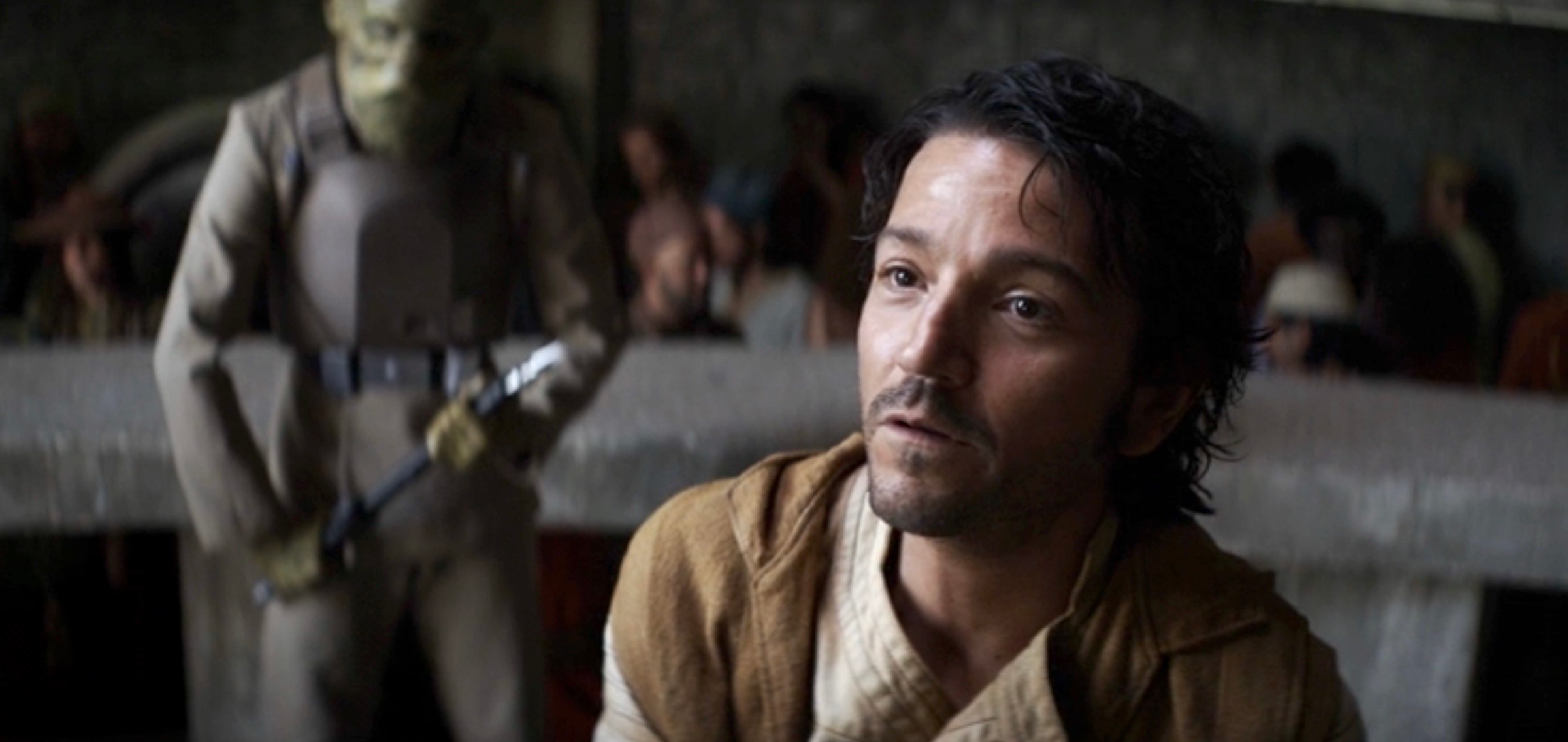 Diego Luna as Cassian Andor pretending to be Keef Girgo, wearing a tan vest as he pleads at the court bench, while a green alien guard stands behind him with a weapon in hand