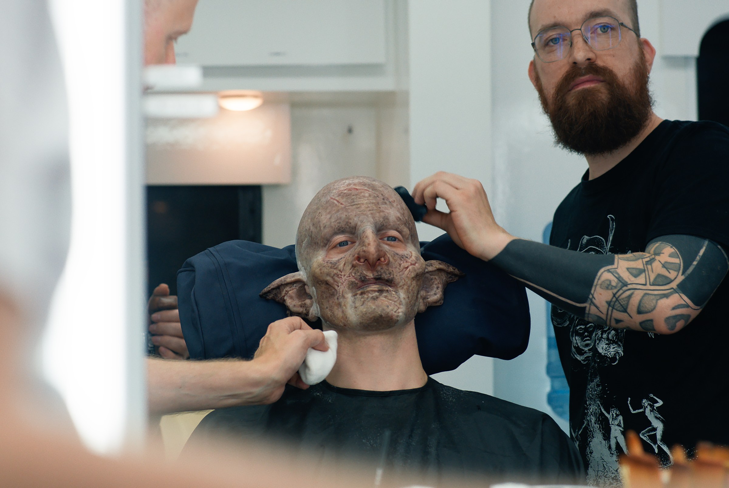 Robert Strange sitting in a chair in partial orc make-up with prosthetics artist Patt Foad tending to something on his head