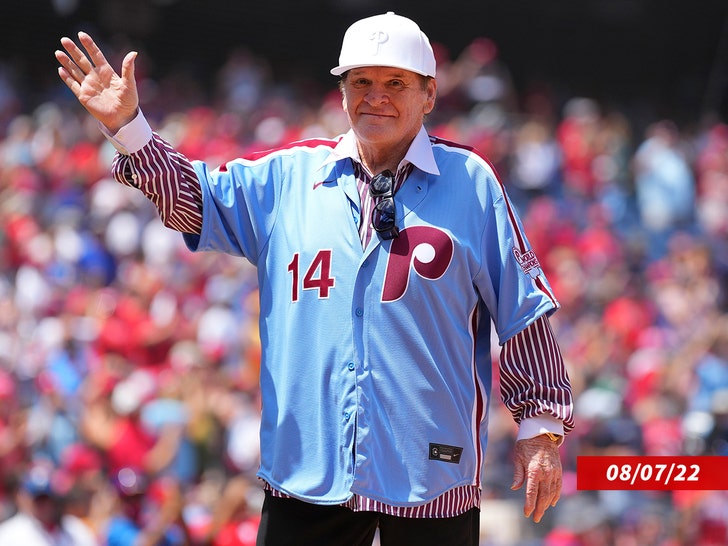 Pete Rose waving while wearing a pin striped shirt under a blue Philadelphia Phillies jersey and white Phillies hat.