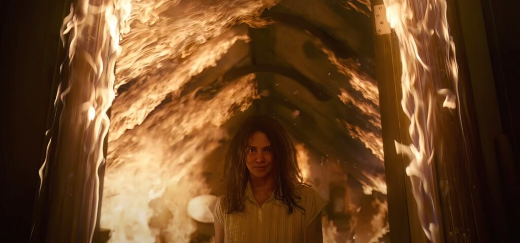 Mama (Halle Berry), a smiling woman in a white dress, stands under a burning roof in Never Let Go