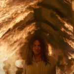 Mama (Halle Berry), a smiling woman in a white dress, stands under a burning roof in Never Let Go