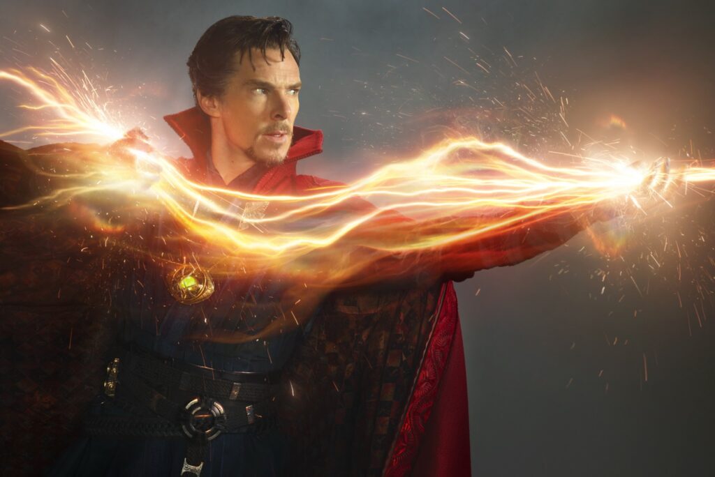 Benedict Cumberbatch as Doctor Strange, in a publicity photo showing him in front of a neutral background, zapping something offscreen with a huge wave of bright orange magical energy