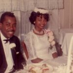 A couple on their wedding day sits at a table and smiles.