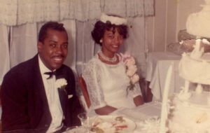 A couple on their wedding day sits at a table and smiles.