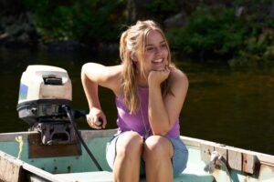 Confident, blonde teenage girl Elliott (Maisy Stella) sits on a small motorboat and smiles widely in My Old Ass