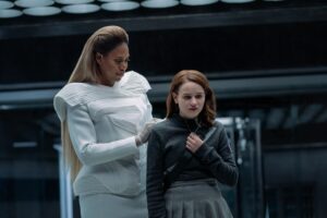 Tally (Joey King), a young woman in grey, looks nervous as a statuesque woman (Laverne Cox) stands behind her in a sterile-looking room in Netflix’s Scott Westerfeld adaptation Uglies