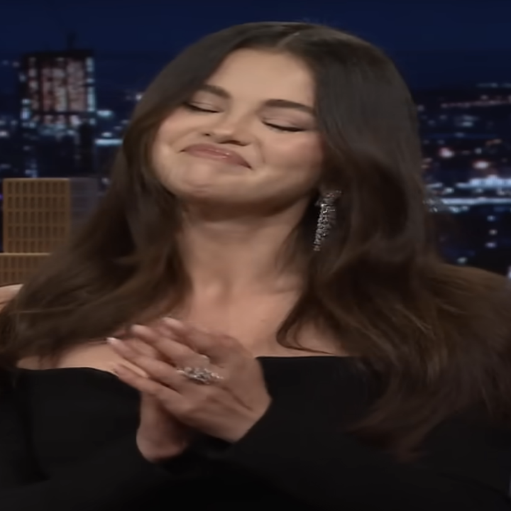 Selena Gomez, wearing an elegant off-shoulder black dress with long, wavy hair, smiling and clapping on a talk show set with a cityscape backdrop