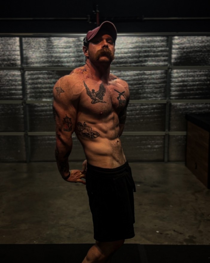 a shirtless man with tattoos on his chest stands in front of a garage door