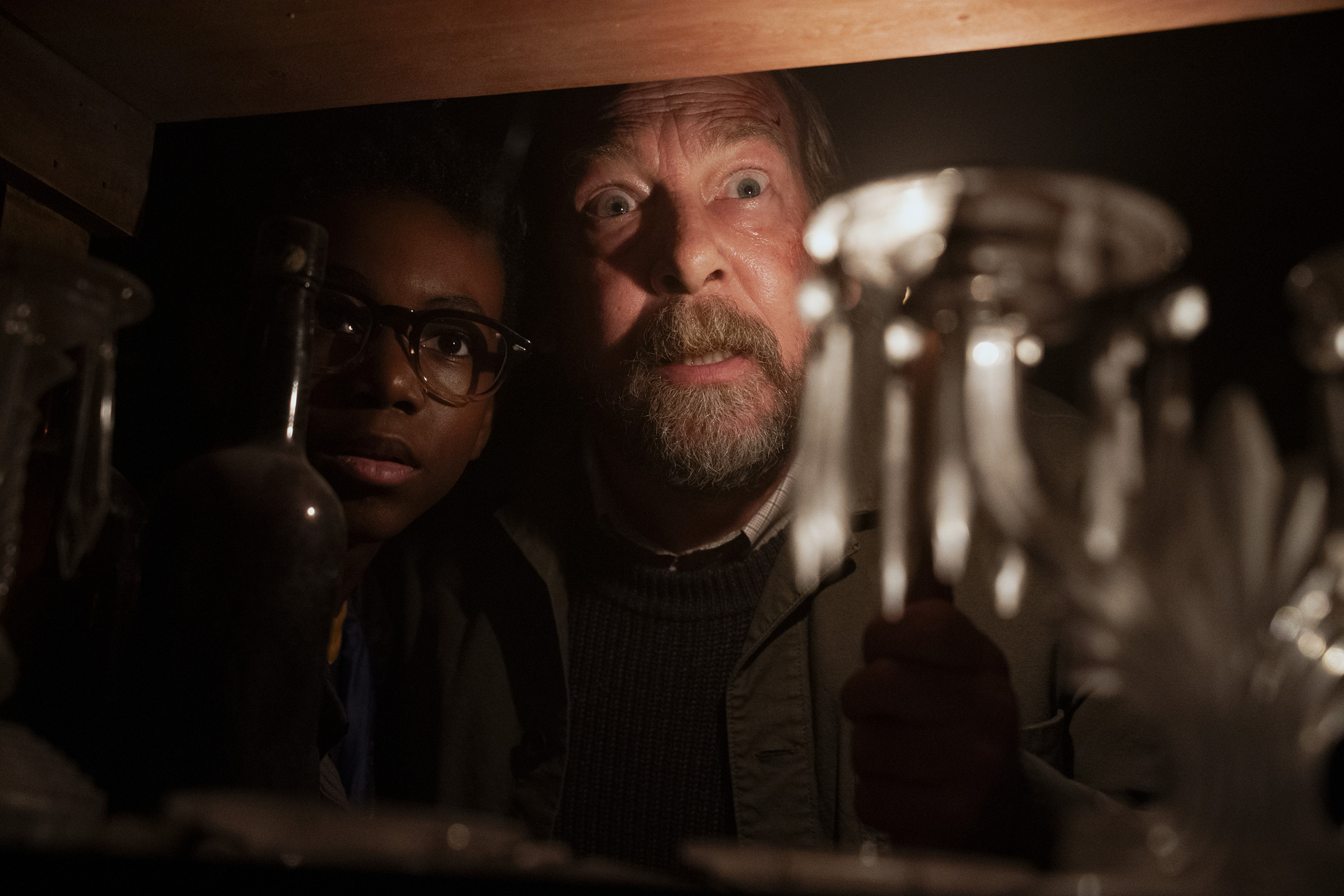 A man with a beard and a teenager wearing glasses look through a shelf of glassware as they are lit with a flashlight in Salem’s Lot  