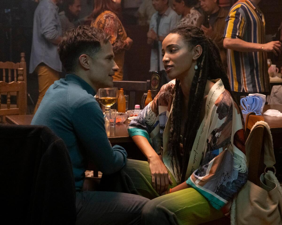 A man in a blue turtleneck sits with a woman with long hair in a loose top in a crowded restaurant.