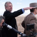 Christopher Nolan's hair is blown by the wind while directing and surrounded by cast and crew on Oppenheimer