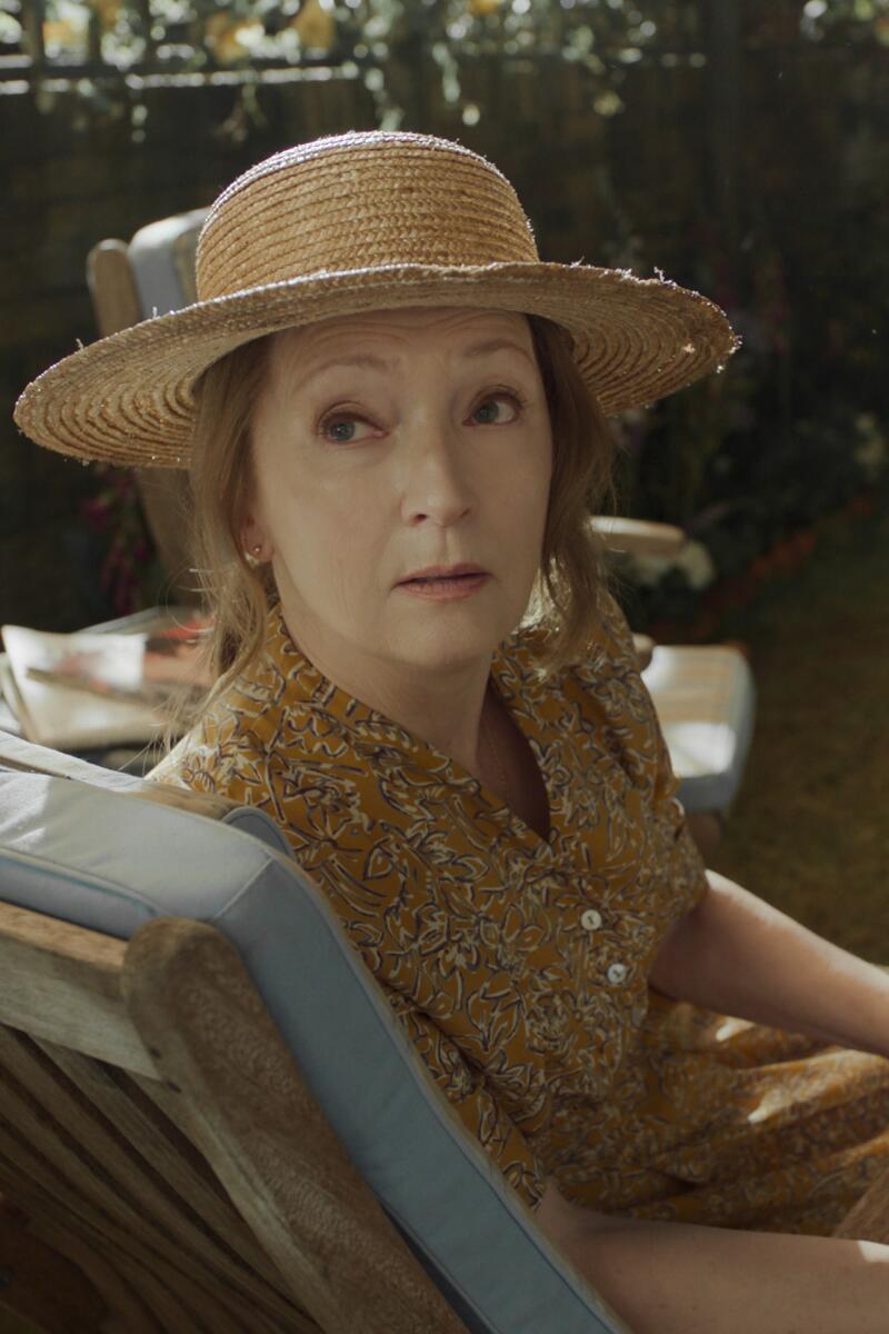 A woman in a sundress and hat sits on a lounge chair and looks over her shoulder.