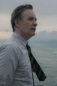 A man in a shirt and tie, seen from mid-waist up, stands with his back to the ocean.