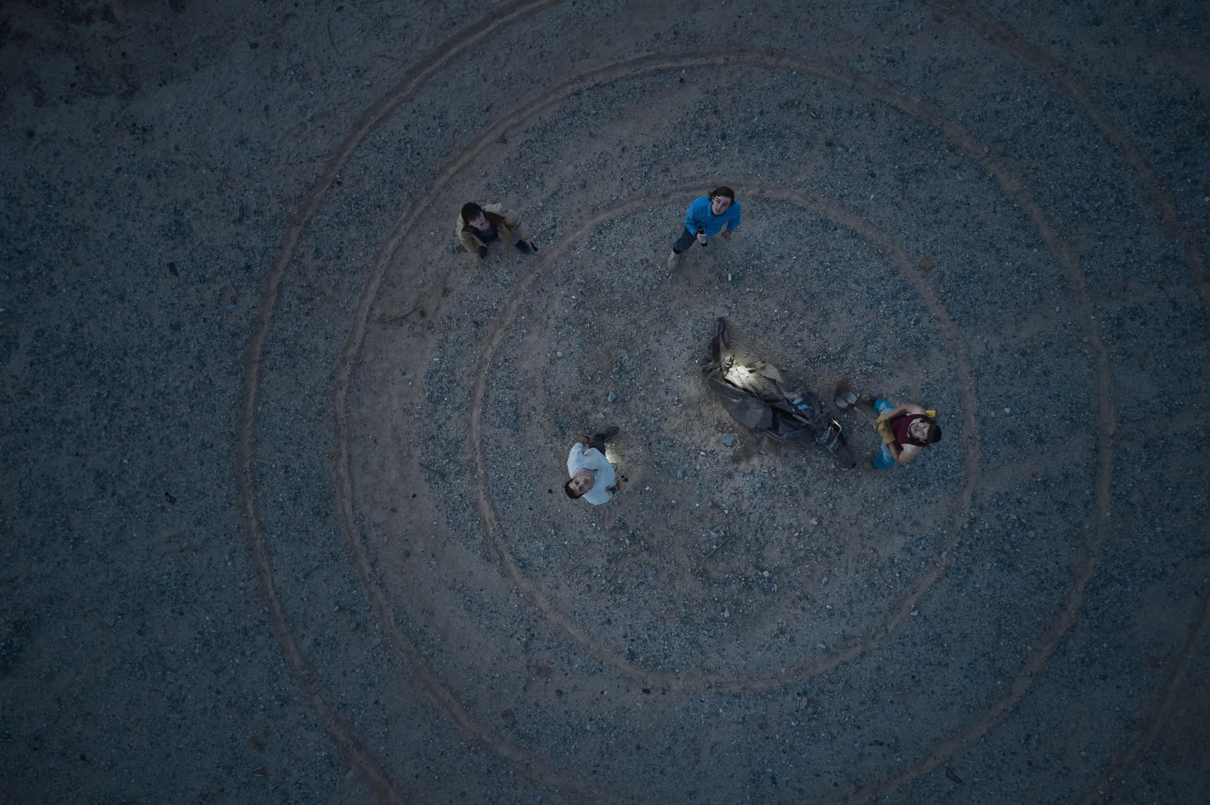 An overhead shot from Falling Stars of four young men standing around an excavated body in the desert. They look up toward the camera. They’re surrounded by circle lines drawn in the dust.