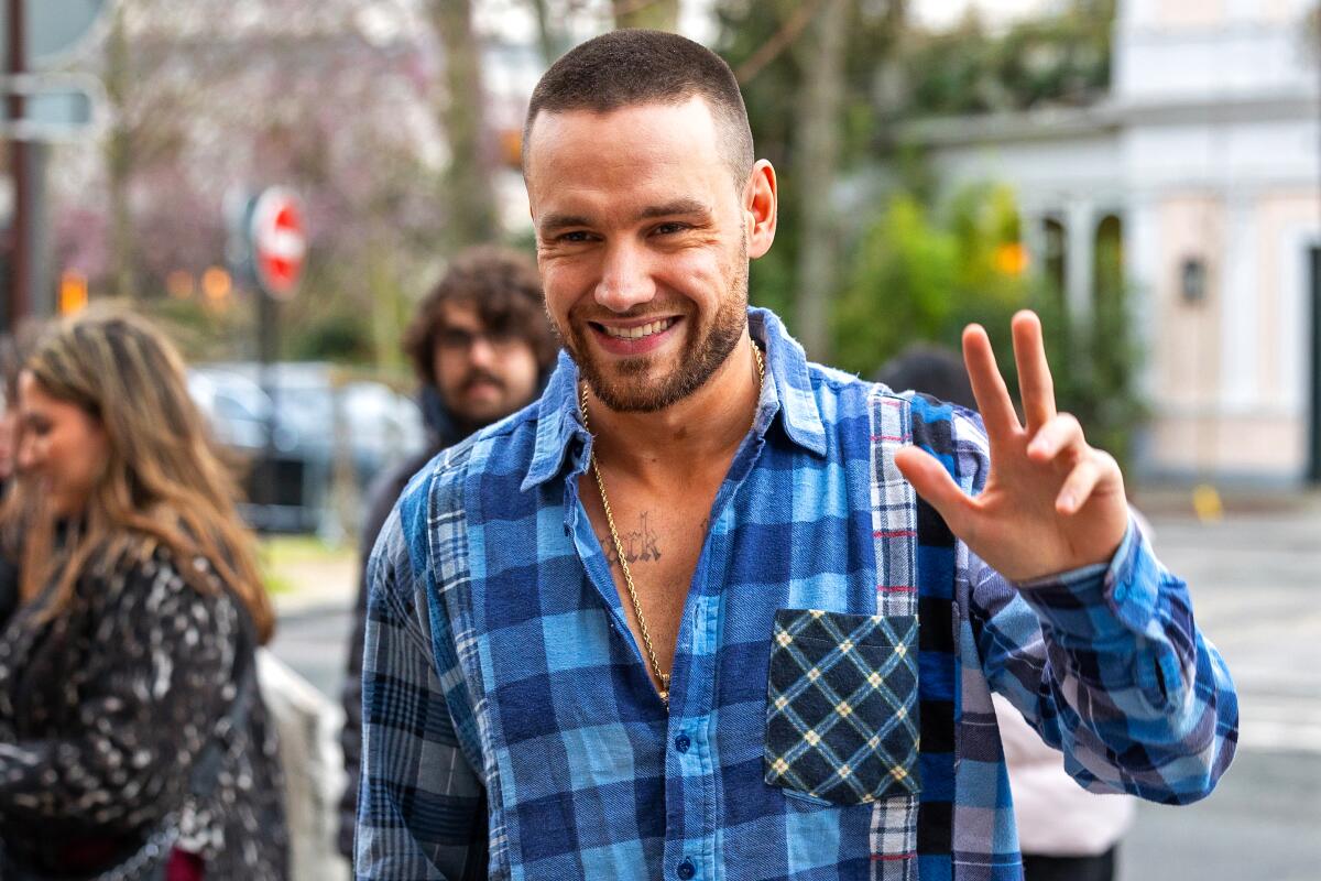 Liam Payne with a shaved head wearing a blue flannel shirt smiling and posing with his right hand up