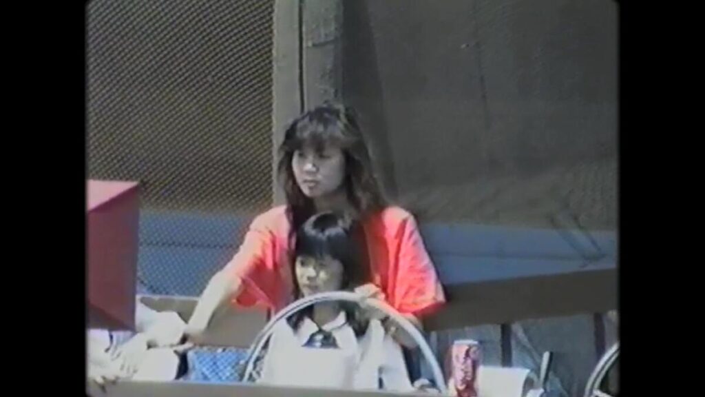 A girl and an older woman stand behind a steering wheel.