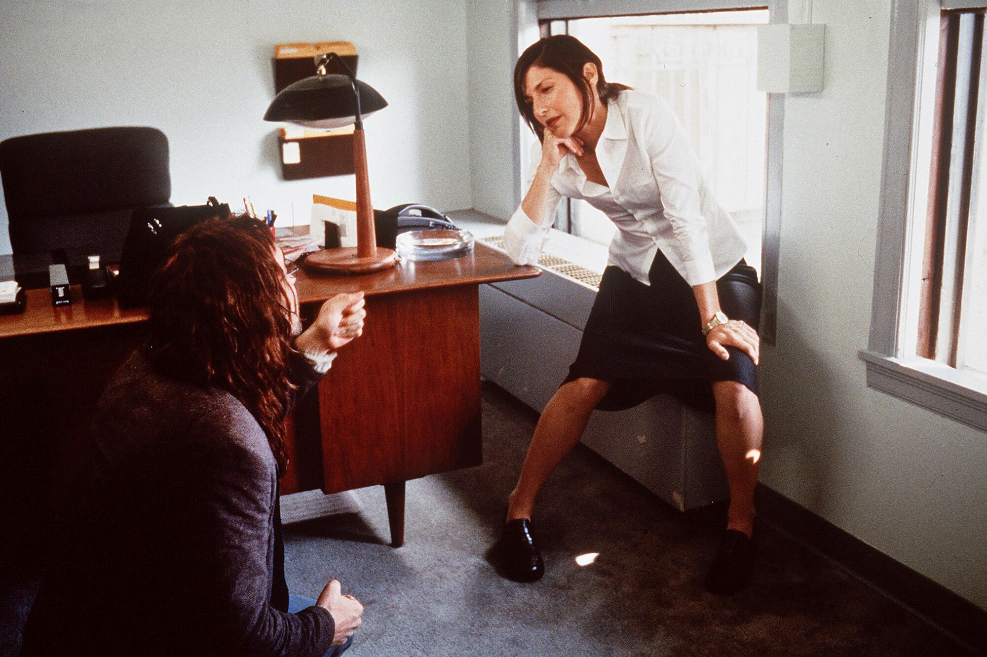 A woman sits on a below-window radiator listening to a seated man talking, in an office.