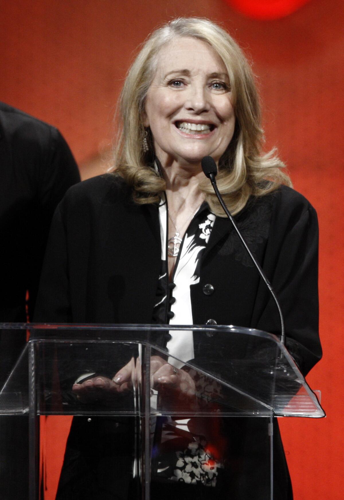 Teri Garr in a black blazer and a white blouse standing on a stage behind a clear podium against a red background