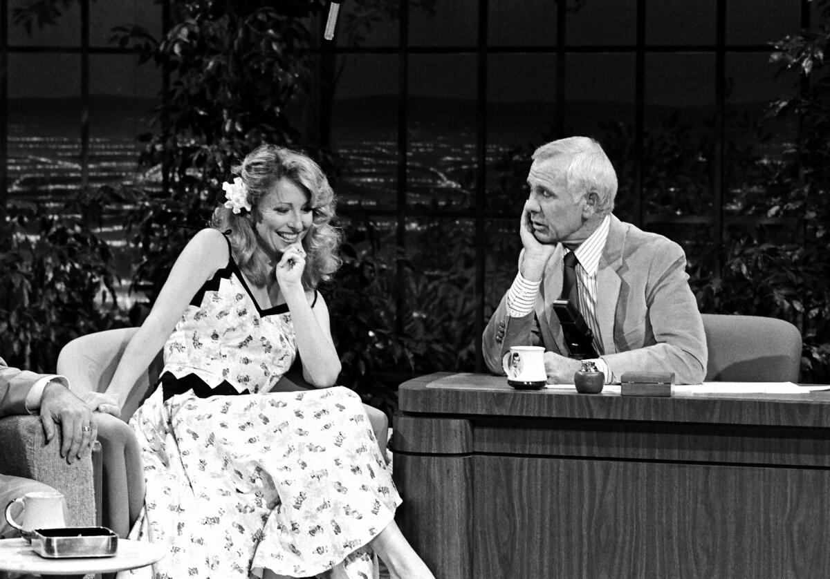 Teri Garr sitting and holding her hand to her chin while talking with Johnny Carson who is seated behind a desk