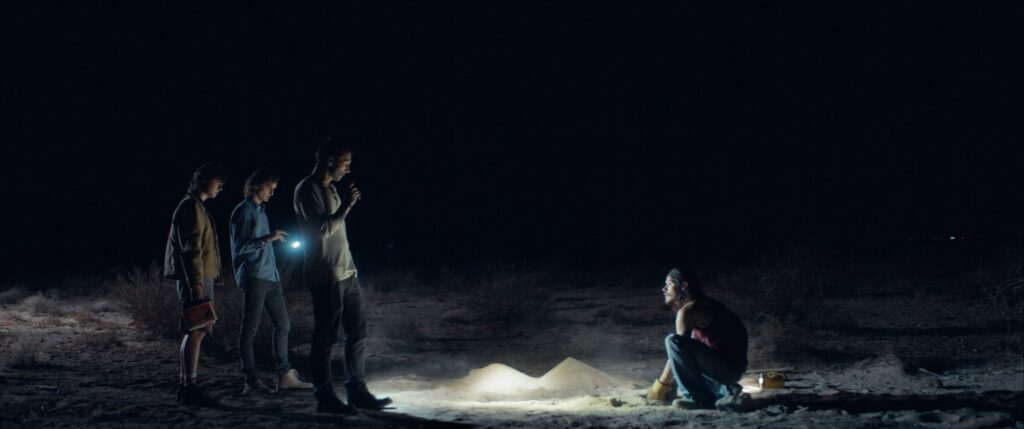 A side shot of four men around an excavated grave in the desert at night, in Falling Stars. Three look on holding torches while the other kneels down