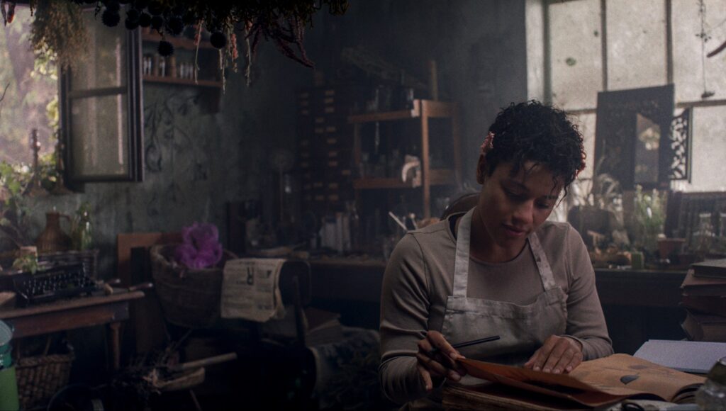 Chef (Ariana DeBose) peruses sheets of parchment and paper in a dark, overgrown, cluttered manor-house kitchen in House of Spoils