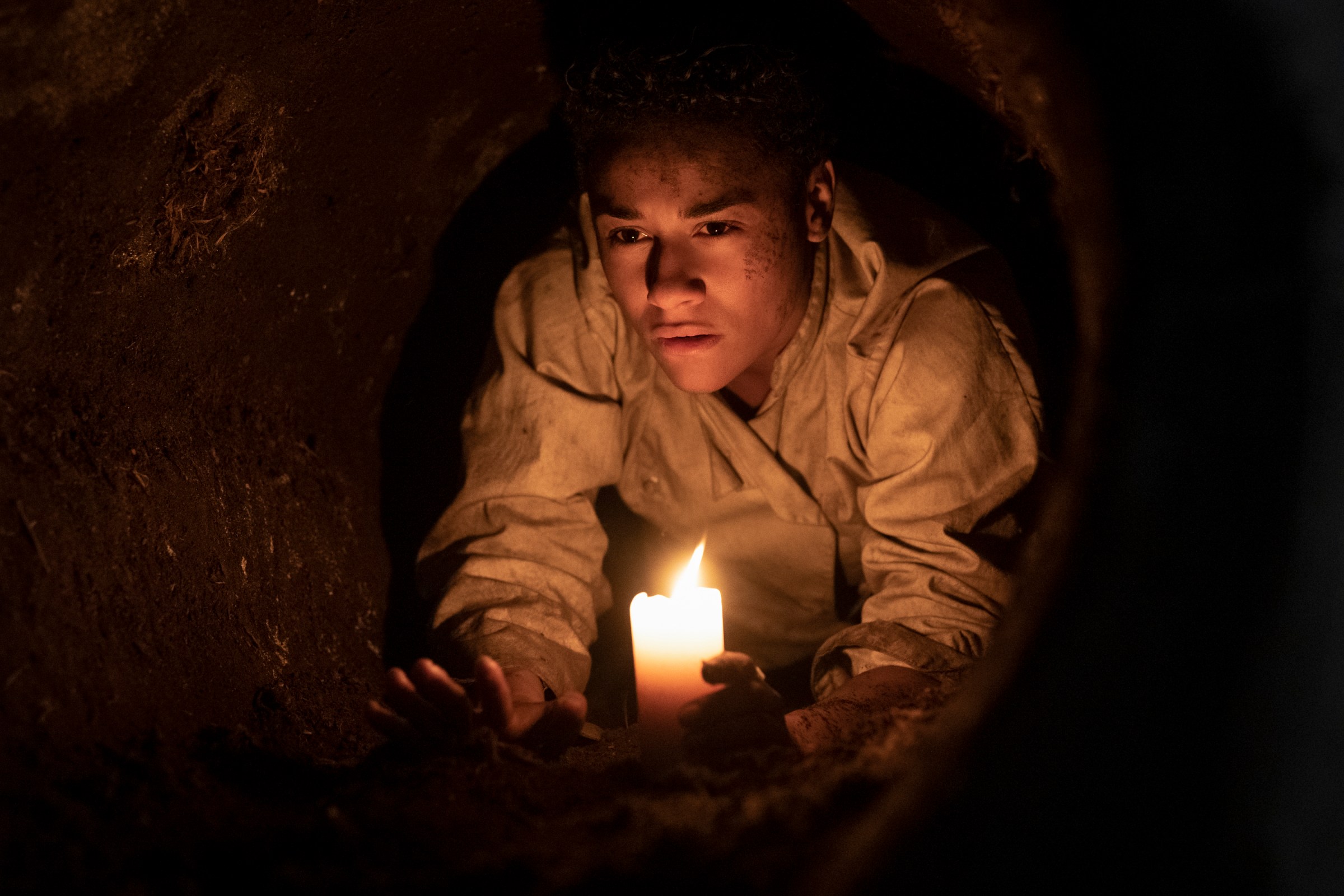 Chef (Ariana DeBose), clutching a candle, crawls through a narrow underground tunnel in House of Spoils