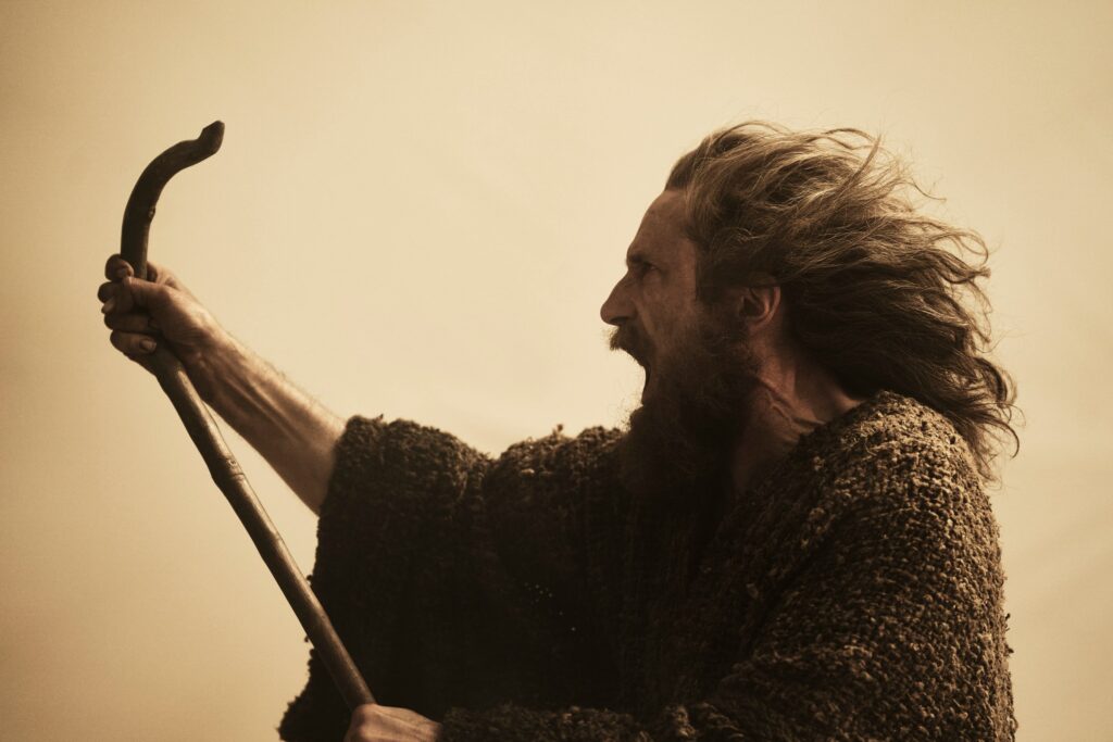 Daniel Weyman as The Stranger in The Lord of the Rings: The Rings of Power. He’s brandishing a staff-like branch into a strong wind and shouting.