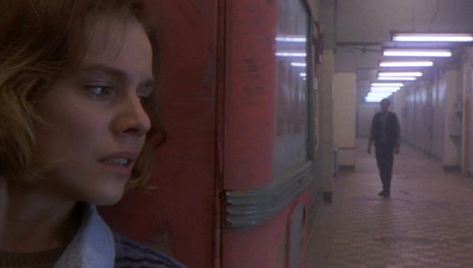 A woman leaning around a corner with a man standing at the end of a long hallway in Mute Witness.